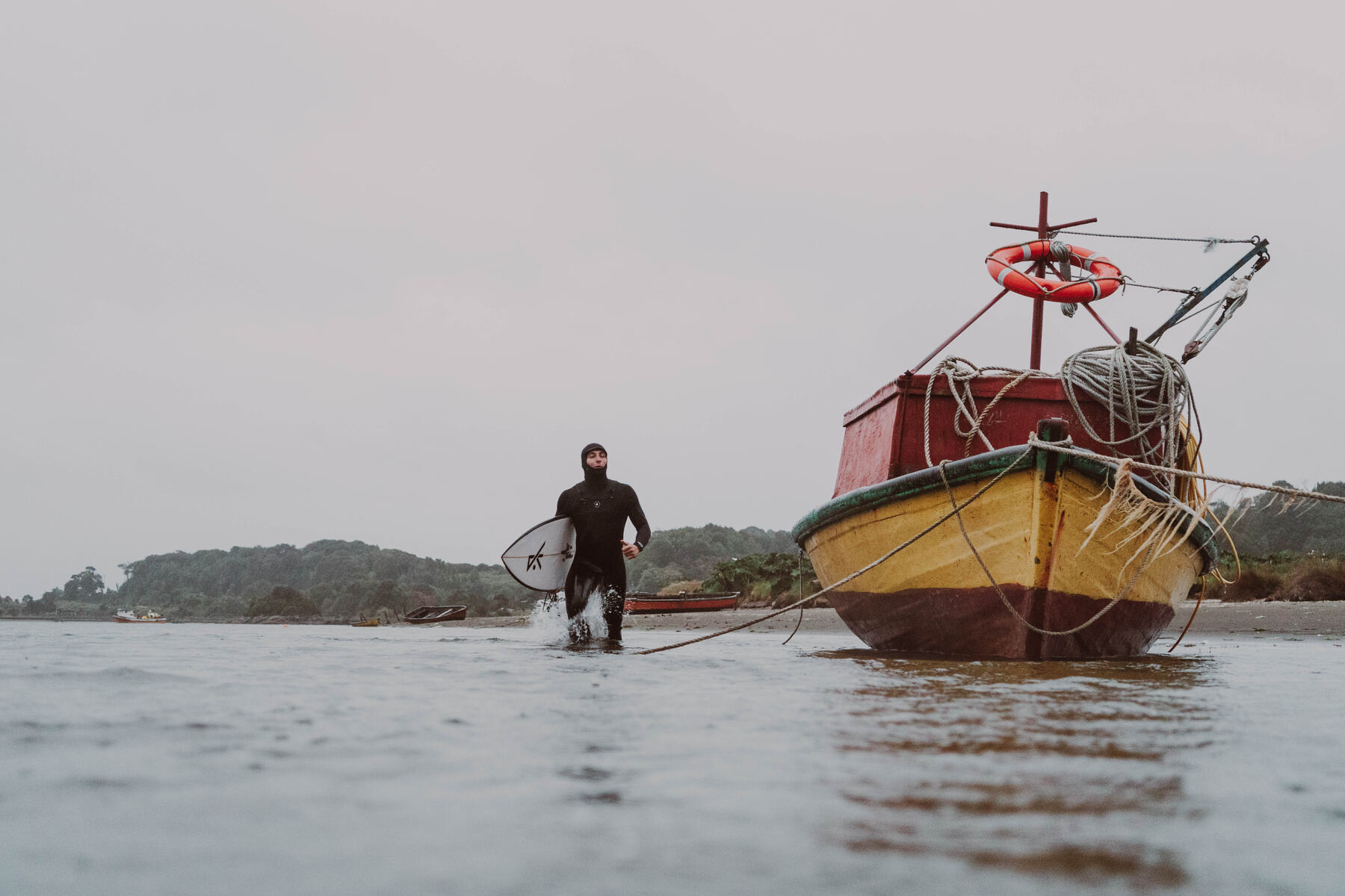 Surfista con su tabla caminando en la orilla junto a un bote de pesca amarillo